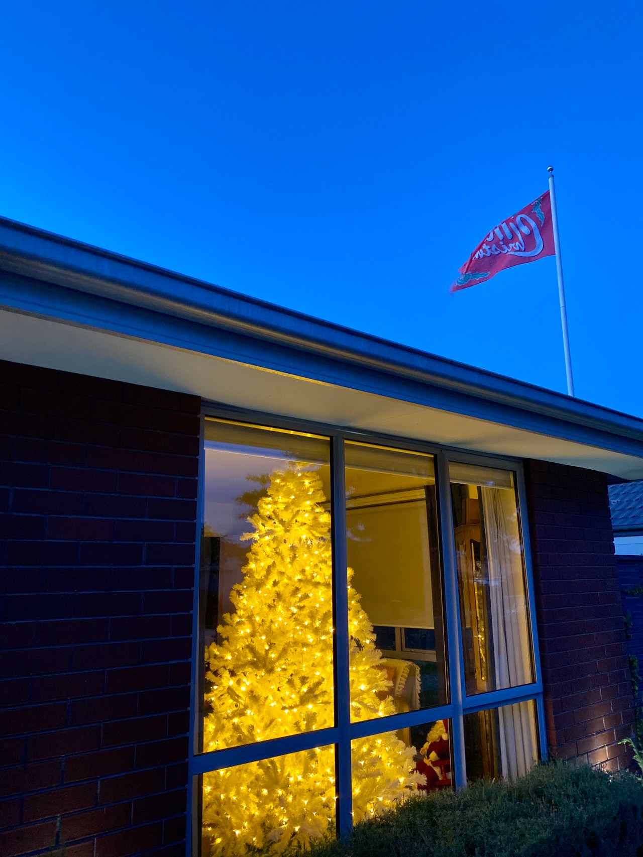 White Christmas tree illuminated with warm white lights in the window of a house with a Merry Christmas flag flying outside as the sun is setting