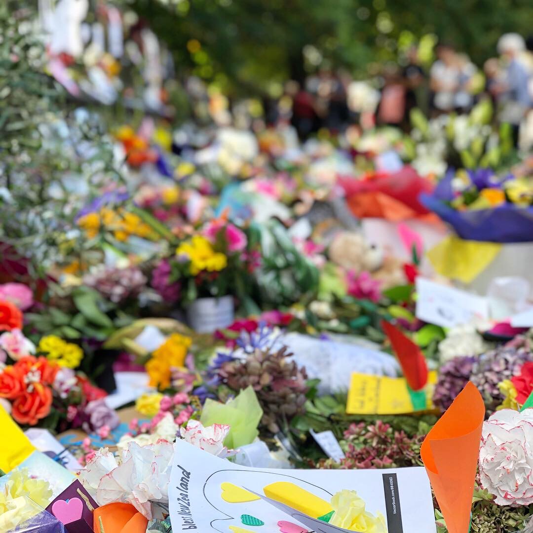 Floral tributes and signs and a chain of paper humans holding hands on a fence in the Christchurch Botanical Gardens. The main sign is an illustration of Prime Minister Jacinda Adern hugging a woman in a hijab with the words, "New Zealand is their home. They are us."