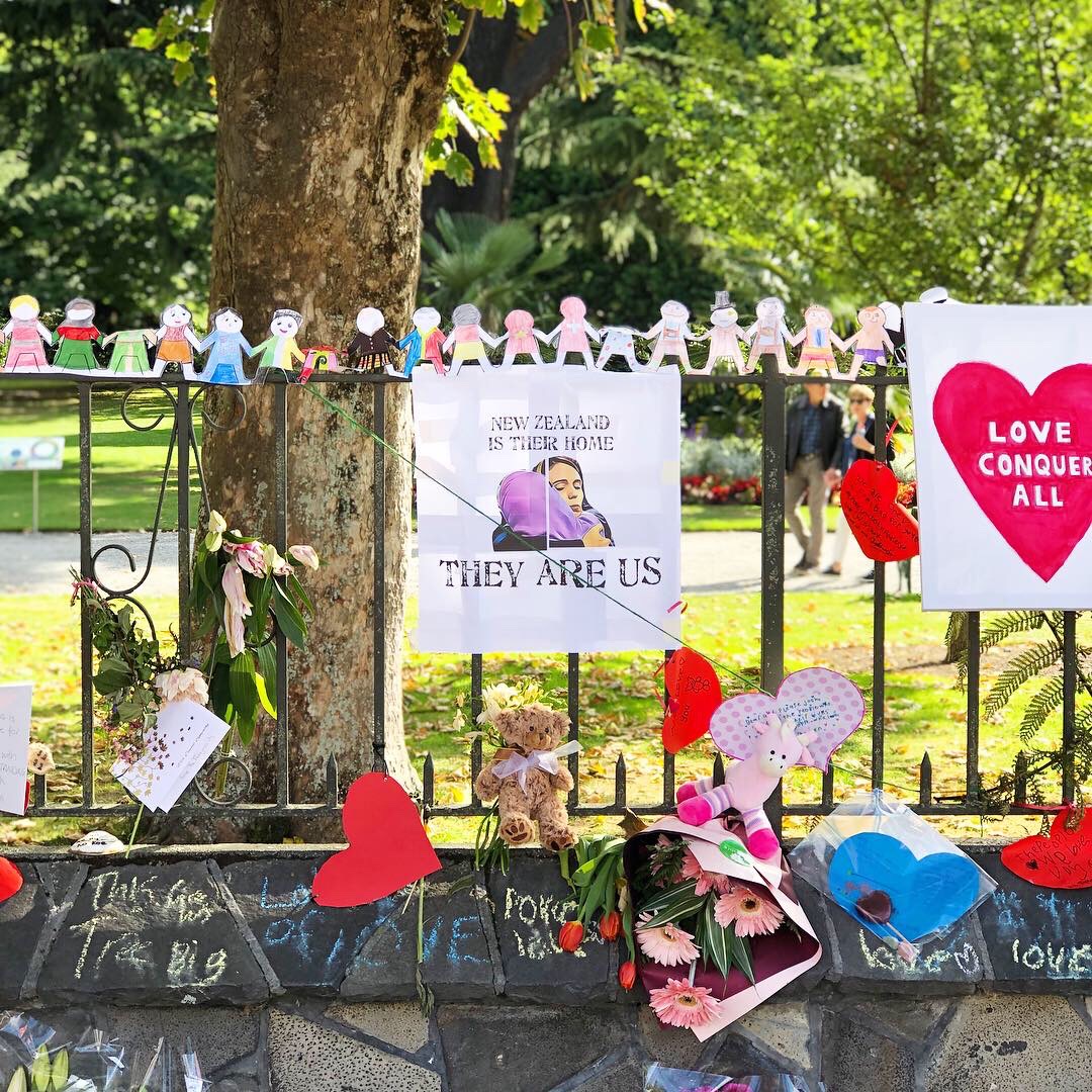 Floral tributes and signs and a chain of paper humans holding hands on a fence in the Christchurch Botanical Gardens. The main sign is an illustration of Prime Minister Jacinda Adern hugging a woman in a hijab with the words, "New Zealand is their home. They are us."