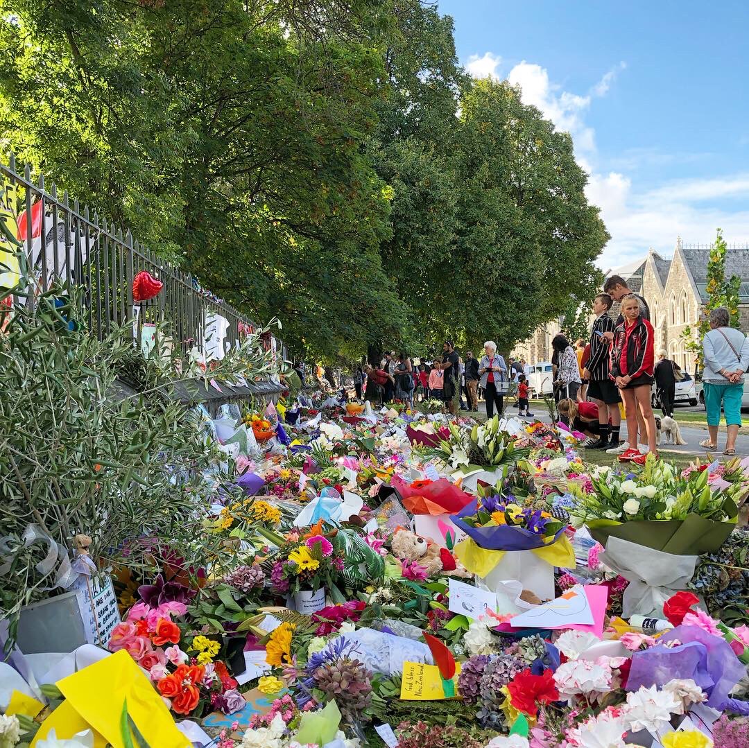 People walk past and pay tribute to the victims of the Christchurch terrorist attacks. There are a deep row of flowers as far as the eye can see pressed up against the fence of the Christchurch Botanical Gardens.