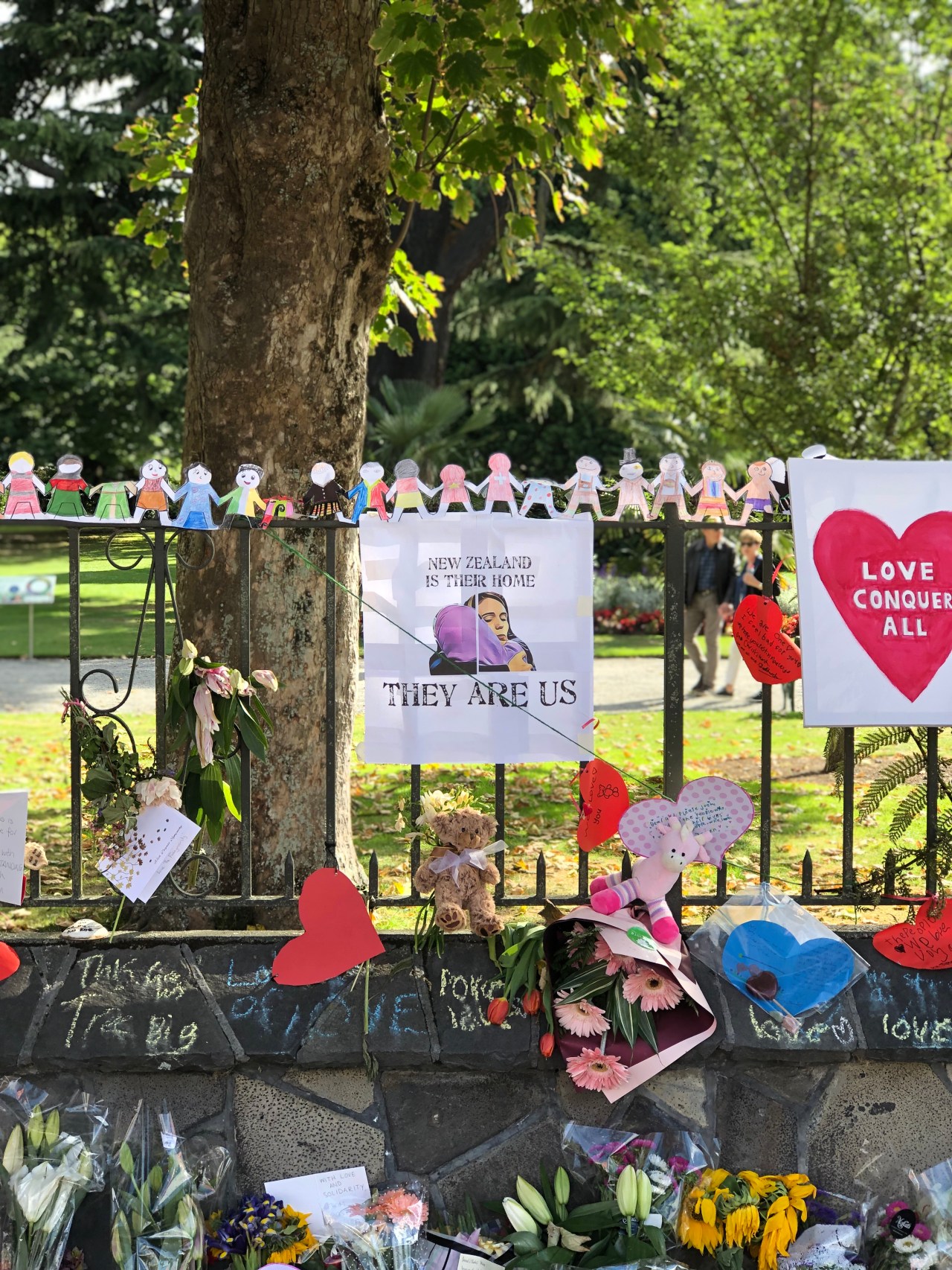 Floral tributes and signs and a chain of paper humans holding hands on a fence in the Christchurch Botanical Gardens. The main sign is an illustration of Prime Minister Jacinda Adern hugging a woman in a hijab with the words, "New Zealand is their home. They are us."