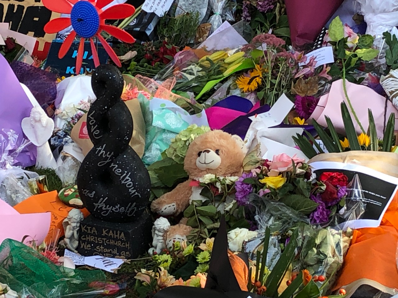 Many floral tributes for the victims of the Christchurch terrorist attacks, with small statues of angels and a teddy bear. A Maori carving sits in the middle with the words, "Love thy neighbour and thyself. Kia kaha Christchurch. We stand as one," handwritten on it.