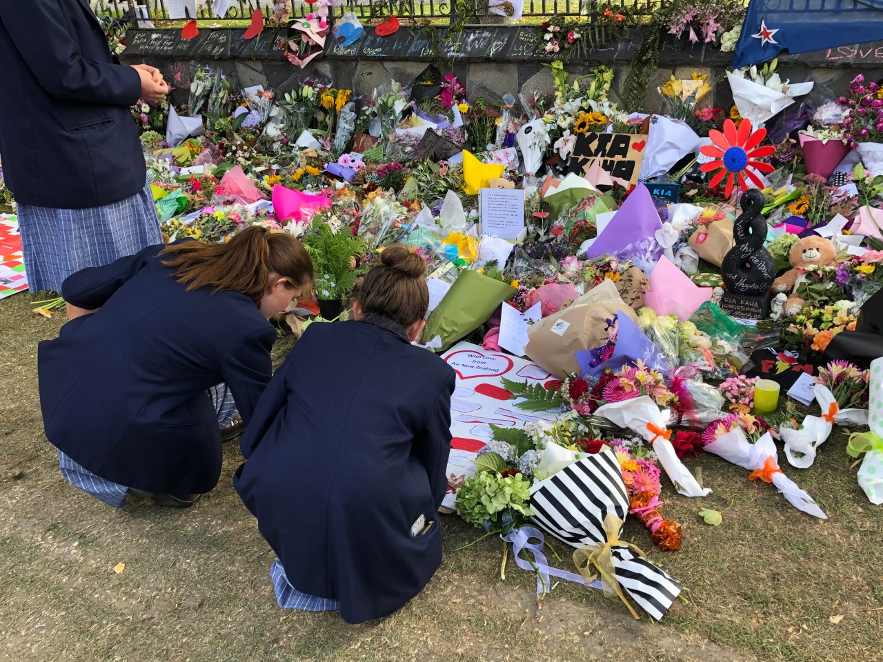 Three high school students write a note on a tribute board surrounded by floral tributes for the victims of the Christchurch terrorist attacks