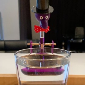 Purple drinking bird in a top hat and red bow tie over a glass of water