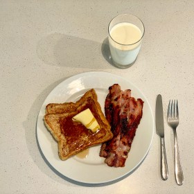 French toast with butter and syrup, and bacon, on a plate, with a knife and fork to the right and glass of milk above it