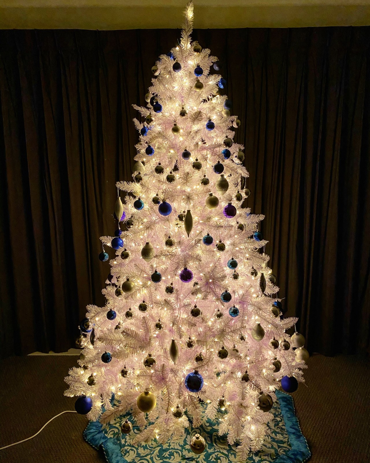 White Christmas tree with blue, teal, and silver ornaments, illuminated with warm white LED lights