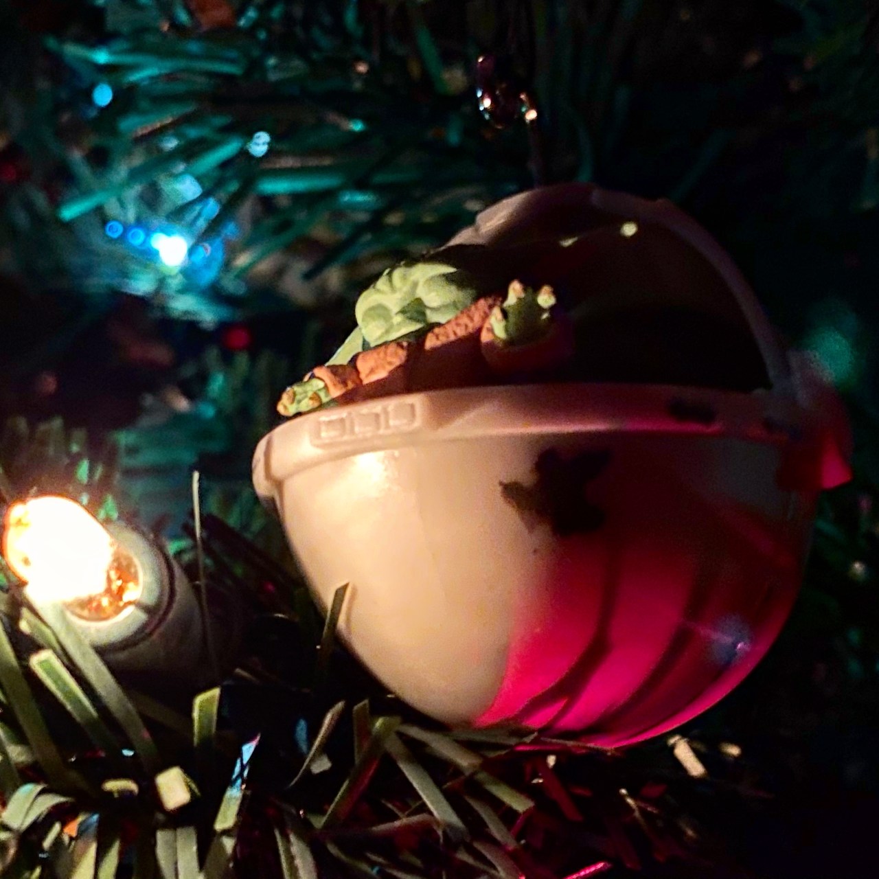 Baby Yoda (Grogu) Christmas ornament. The Child has his eyes closed and his hands up and open, and a yellow Christmas light is in the tree next to him (so it looks like he is levitating it).