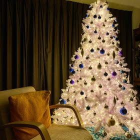 White Christmas tree with white LED lights and blue and silver ornaments, with a reading chair with an orange pillow beside it