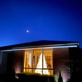 White Christmas tree illuminated in the front window of a house with a twilight darkening blue sky and the moon illuminated in the sky