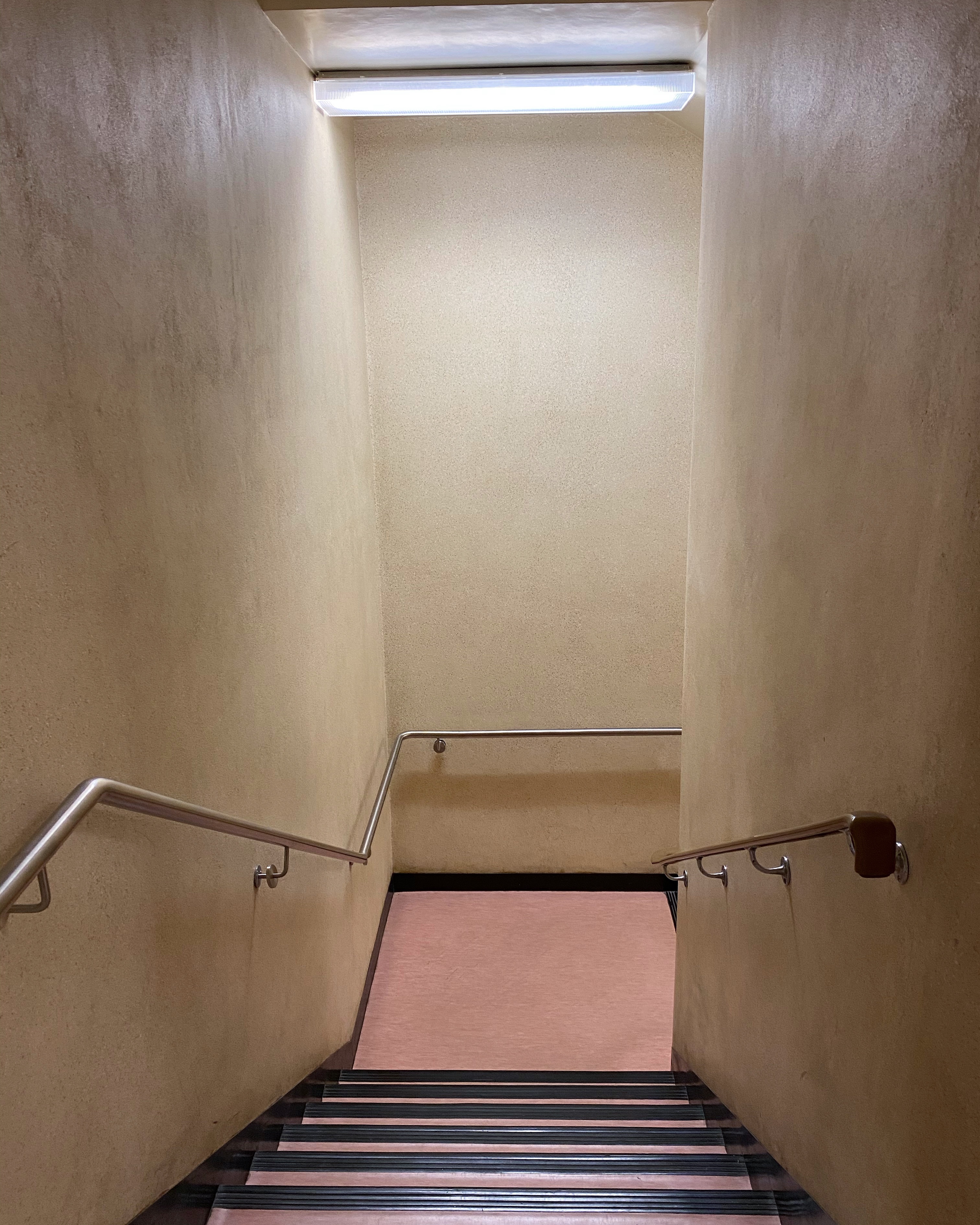 Stairwell heading downstairs, illuminated by a fluorescent light, with dirty butter cream walls and salmon-colored linoleum
