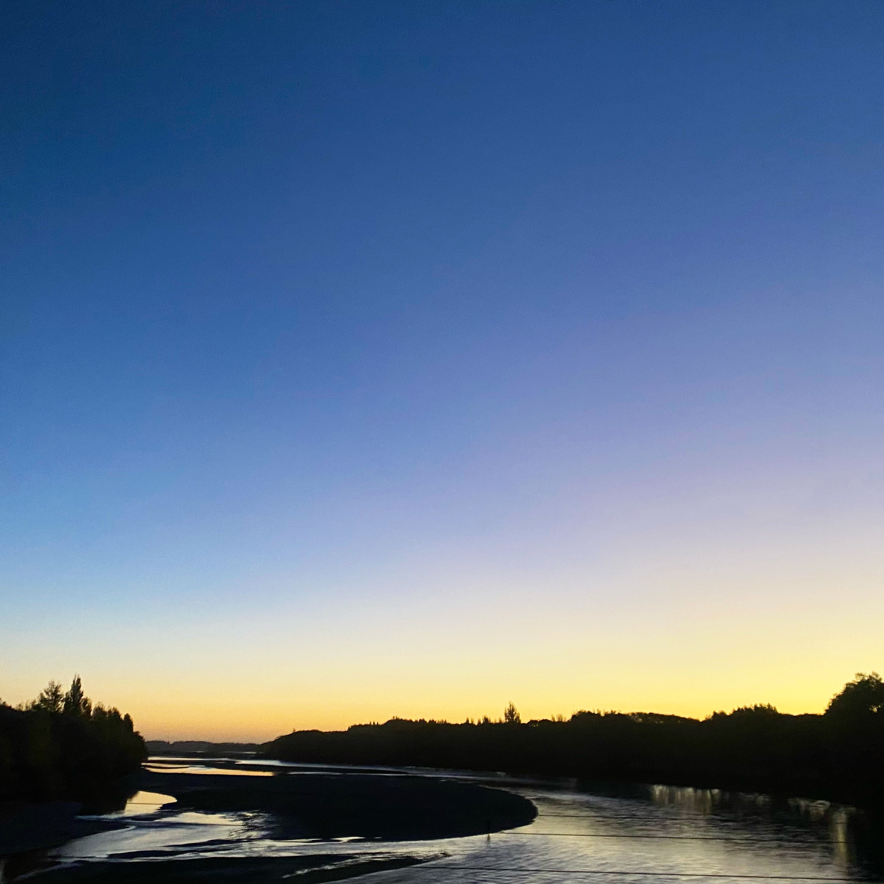 Waimakariri River flowing away from the Southern Alps, with the sun almost set on the horizon. The sky is dark blue at the top to yellow on the horizon.