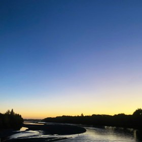 Waimakariri River flowing away from the Southern Alps, with the sun almost set on the horizon. The sky is dark blue at the top to yellow on the horizon.