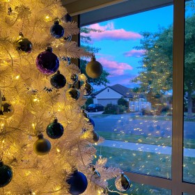 White Christmas tree with warm white LED lights, blue ornaments, and silver ornaments with a darkening blue sky, and pink and purple clouds outside