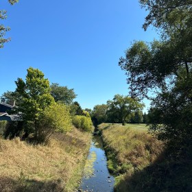 A man-made creek bisects the image of drying grass and green trees with a blue sky above it